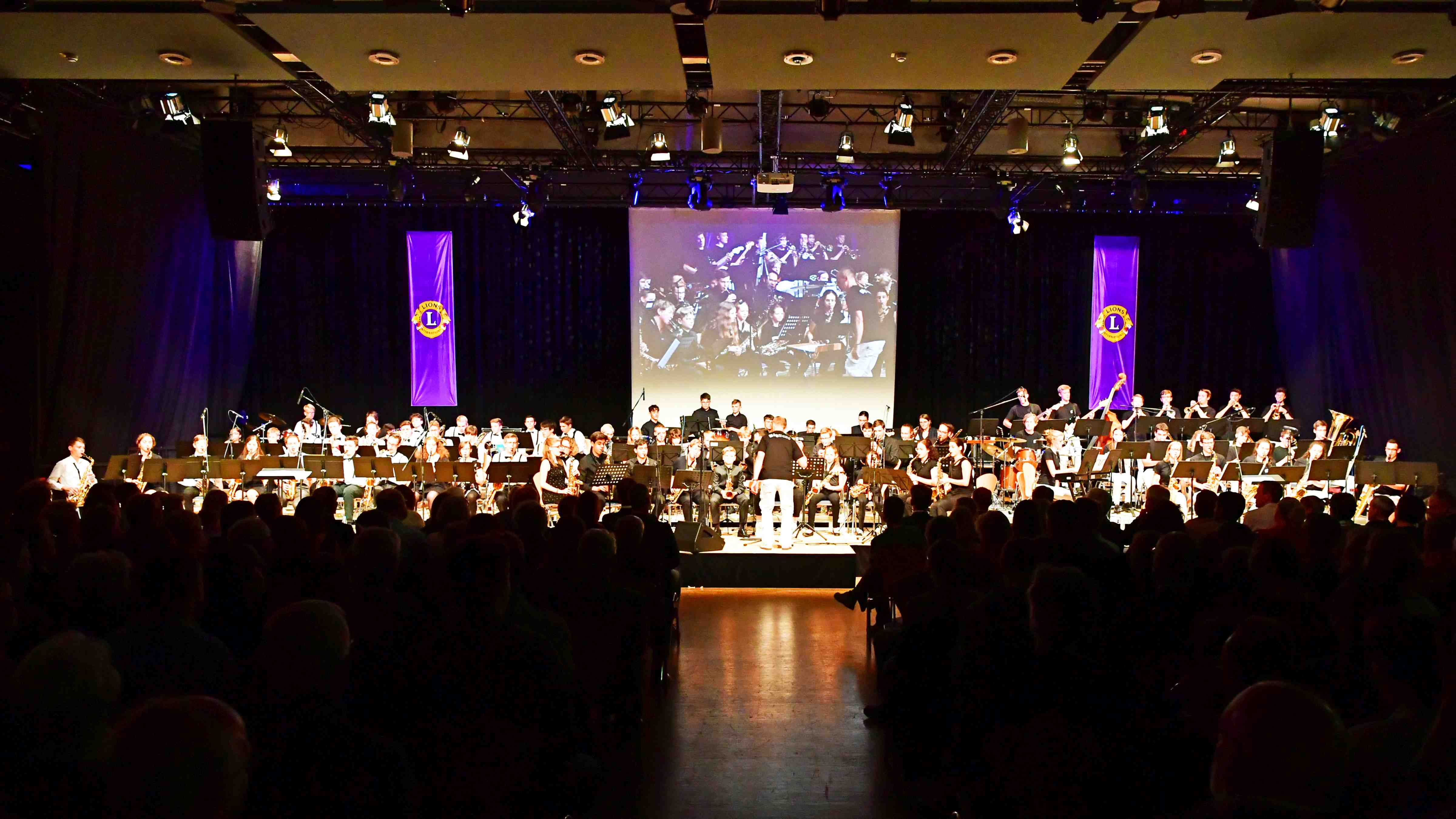Das Foto zeigt die Musiker der Bigbands im Scheinwerferlicht auf der Bühne sowie die Zuhörer auf ihren Stühlen.